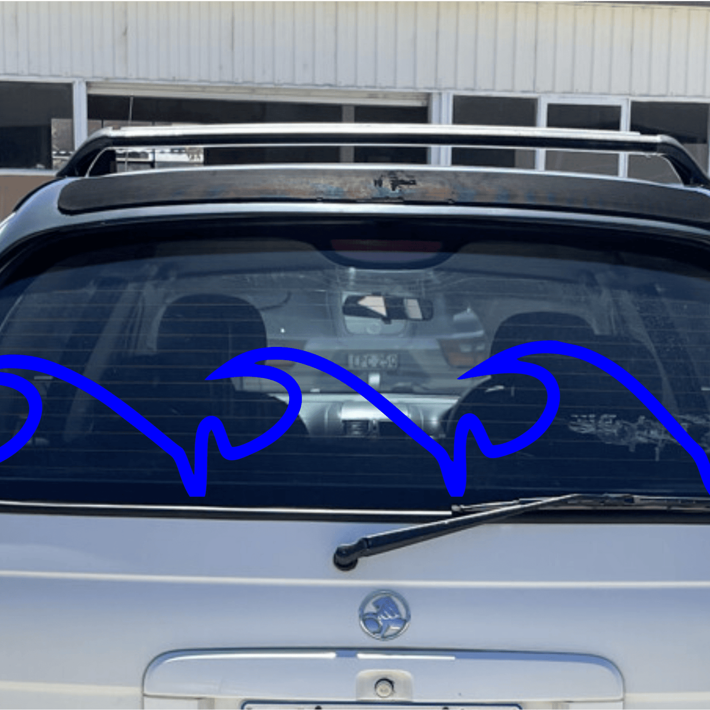 Back of a white car with a blue decorative wave element on the window, parked in front of a building.