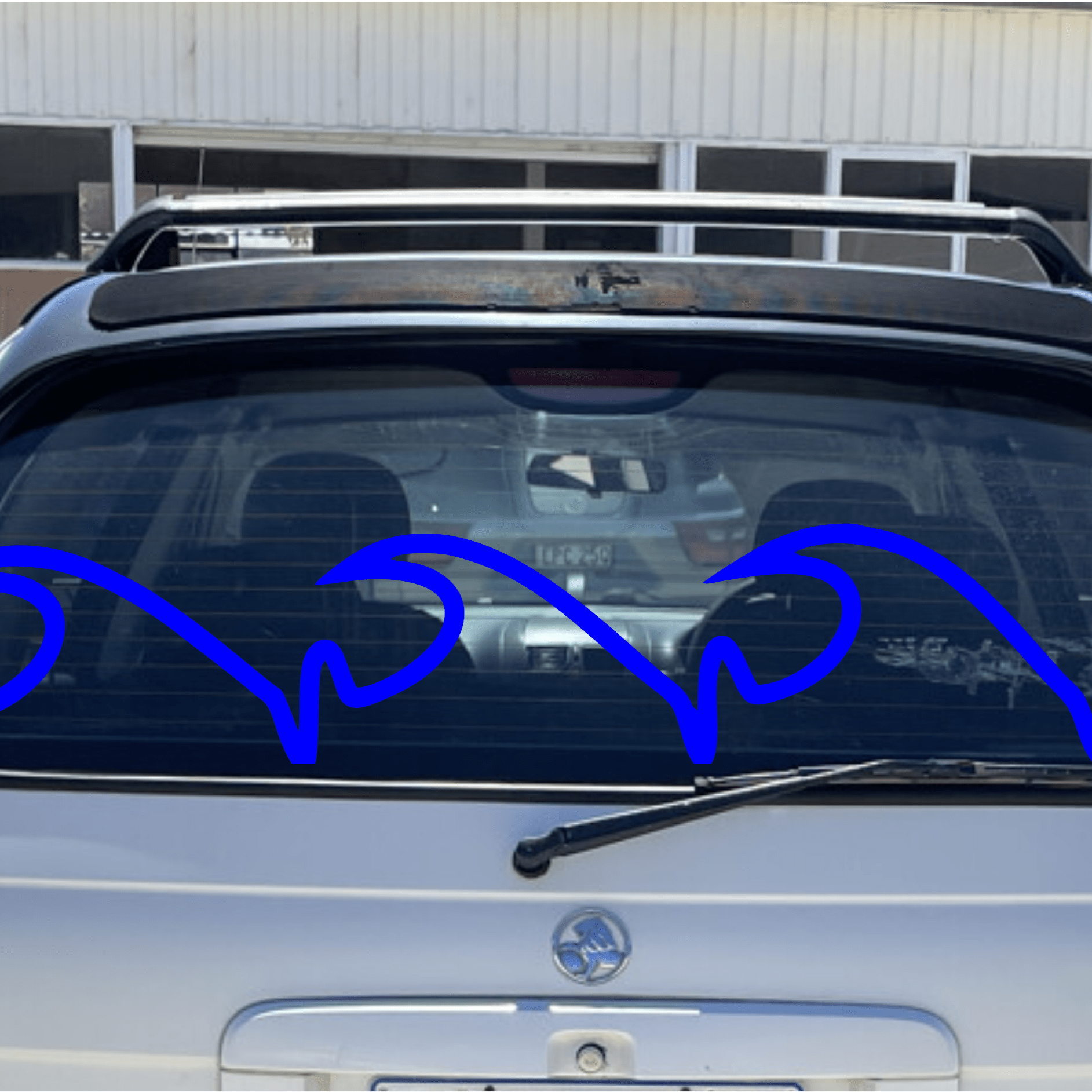 Back of a white car with a blue decorative wave element on the window, parked in front of a building.