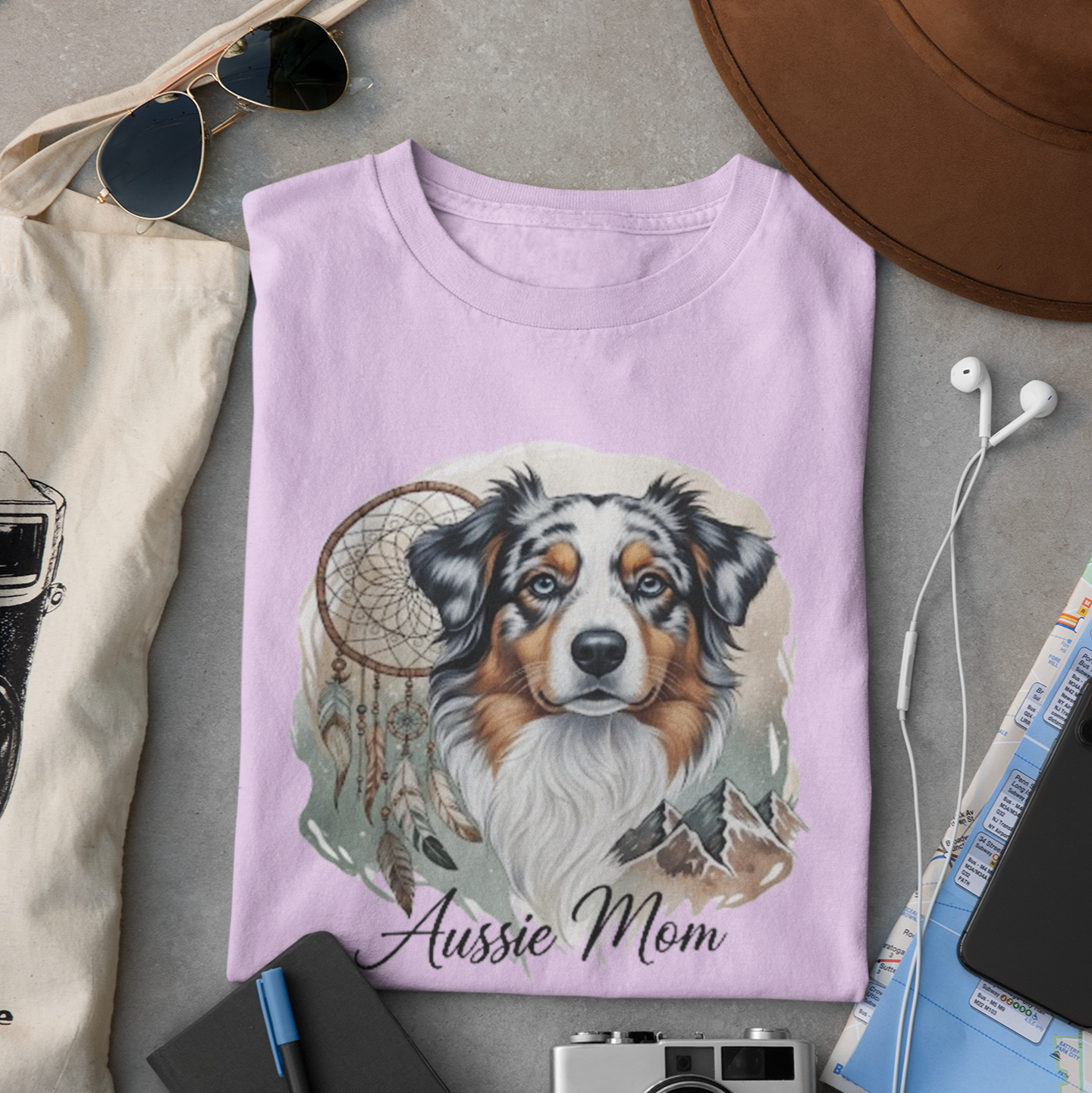 Pink t-shirt with a dog illustration and 'Aussie Mom' text, surrounded by sunglasses, camera, hat, and map on a gray surface.