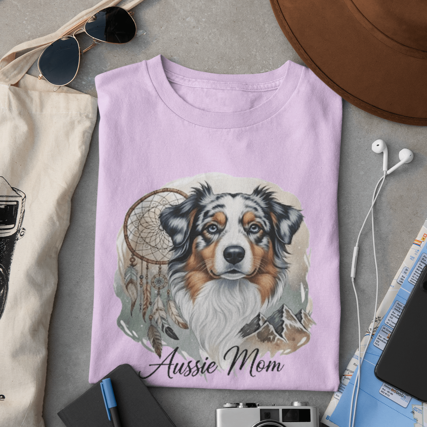 Pink t-shirt with a dog illustration and 'Aussie Mom' text, surrounded by sunglasses, camera, hat, and map on a gray surface.