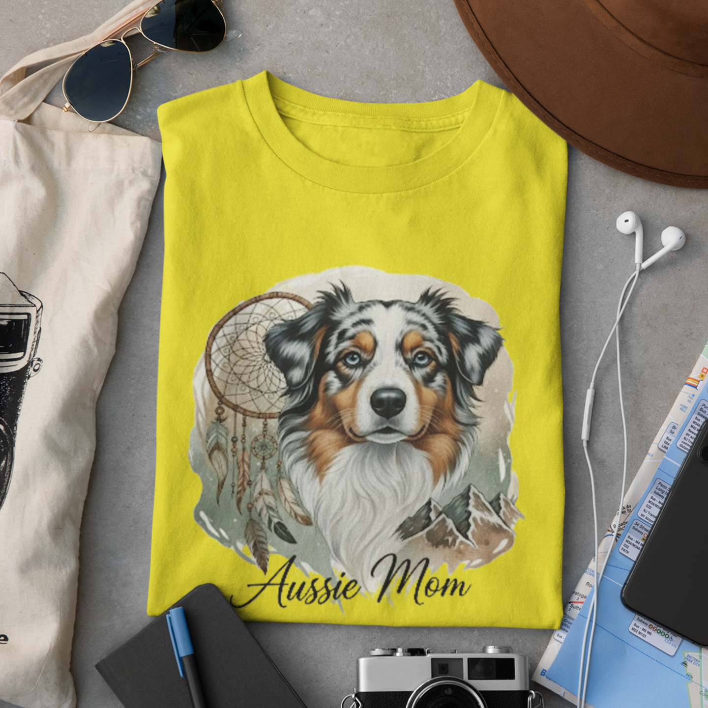 Yellow t-shirt with a dog illustration and 'Aussie Mom' text, surrounded by sunglasses, camera, notebook, and map on a gray surface.