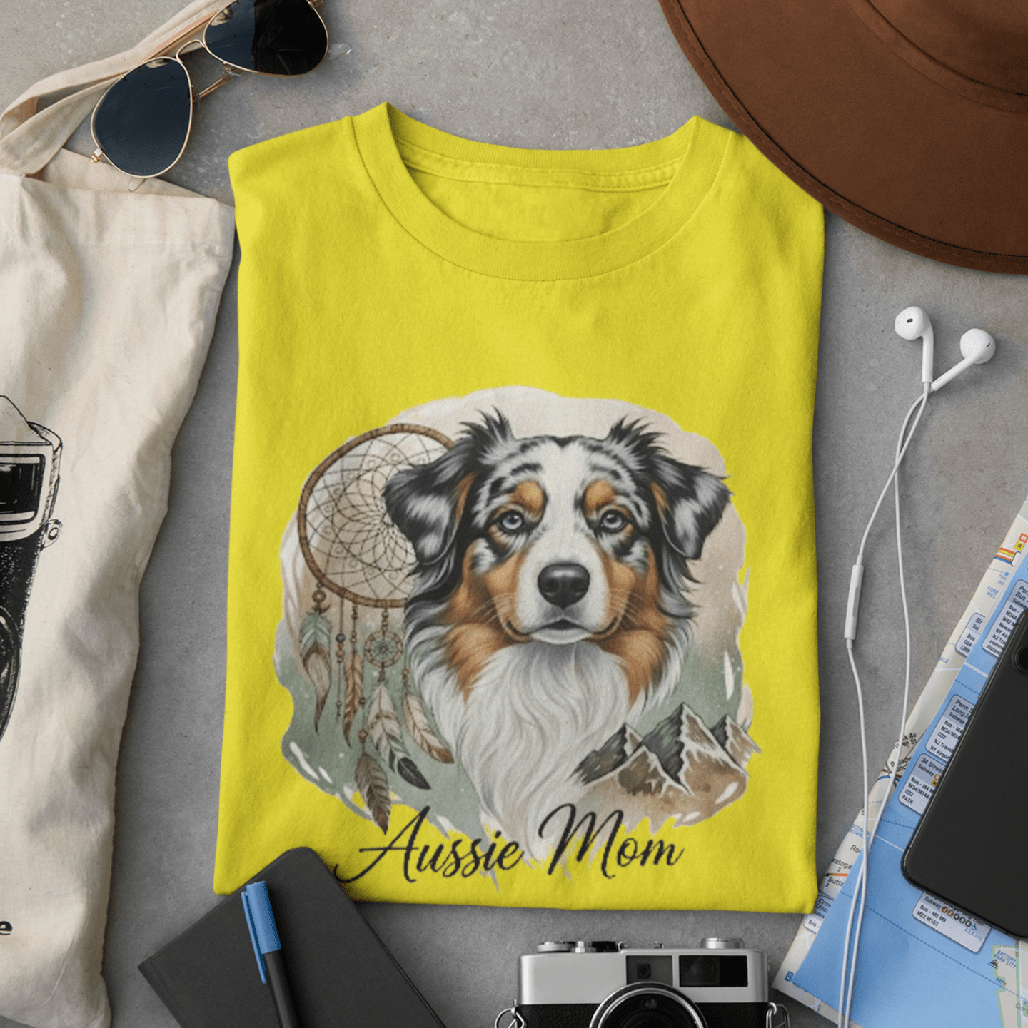 Yellow t-shirt with a dog illustration and 'Aussie Mom' text, surrounded by sunglasses, camera, notebook, and map on a gray surface.