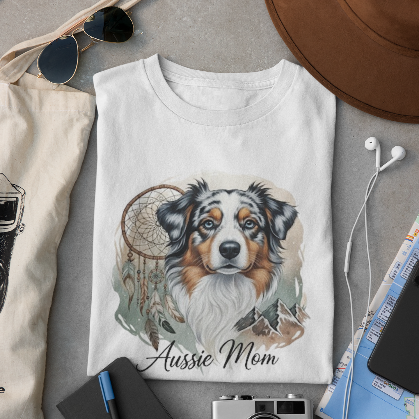 White t-shirt with a dog illustration and 'Aussie Mom' text, surrounded by sunglasses, camera, hat, and notebook on a gray surface.
