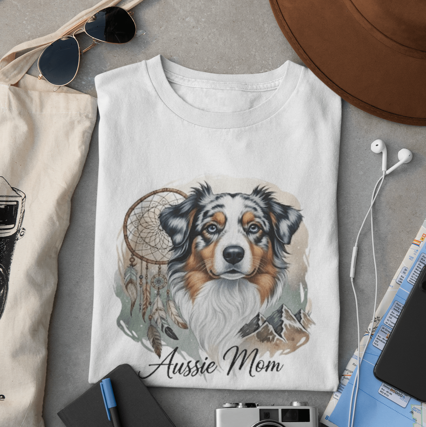White t-shirt with a dog illustration and 'Aussie Mom' text, surrounded by sunglasses, camera, hat, and notebook on a gray surface.