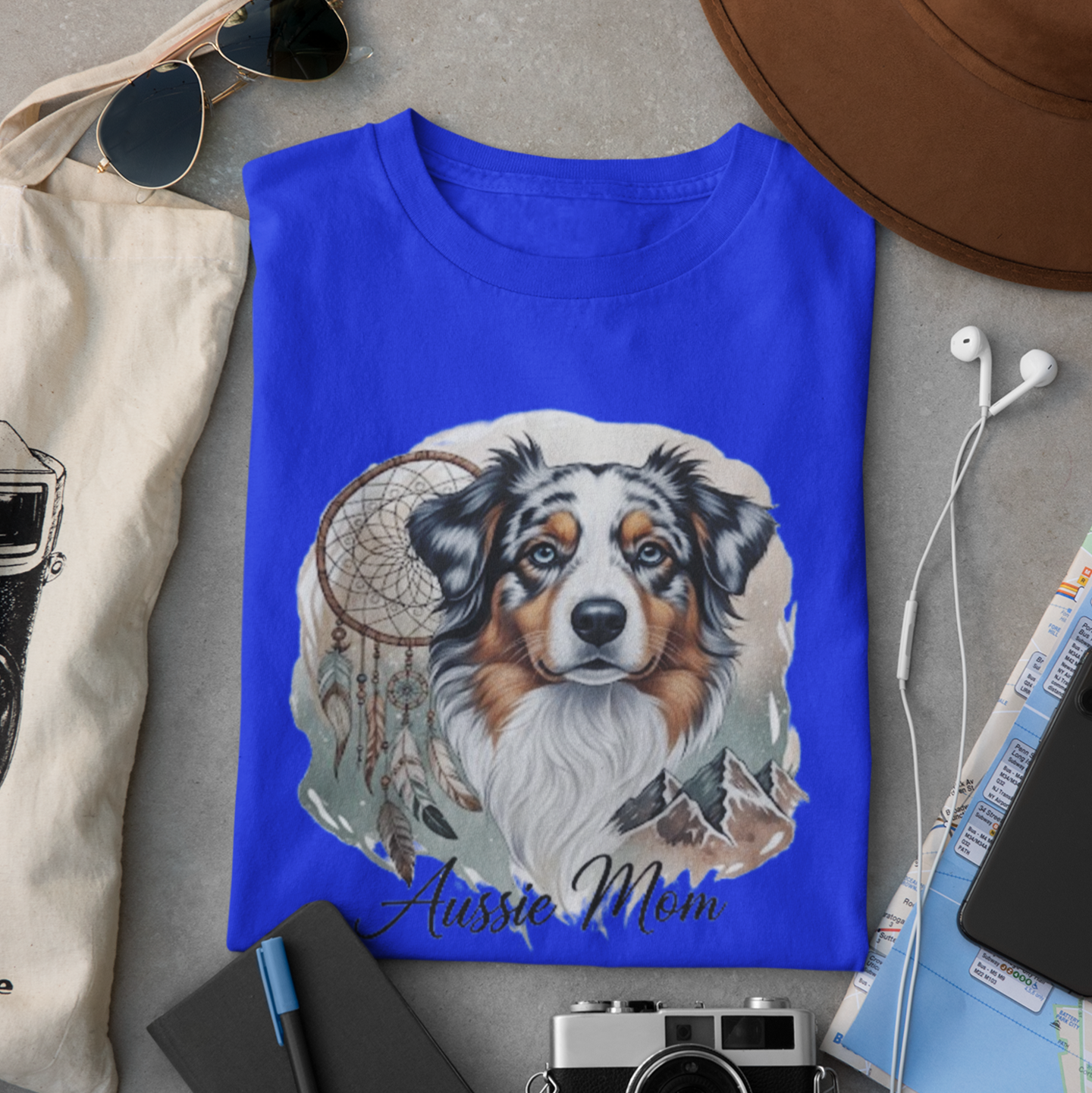 Blue t-shirt with a dog illustration and text, surrounded by sunglasses, camera, hat, and map on a gray surface.