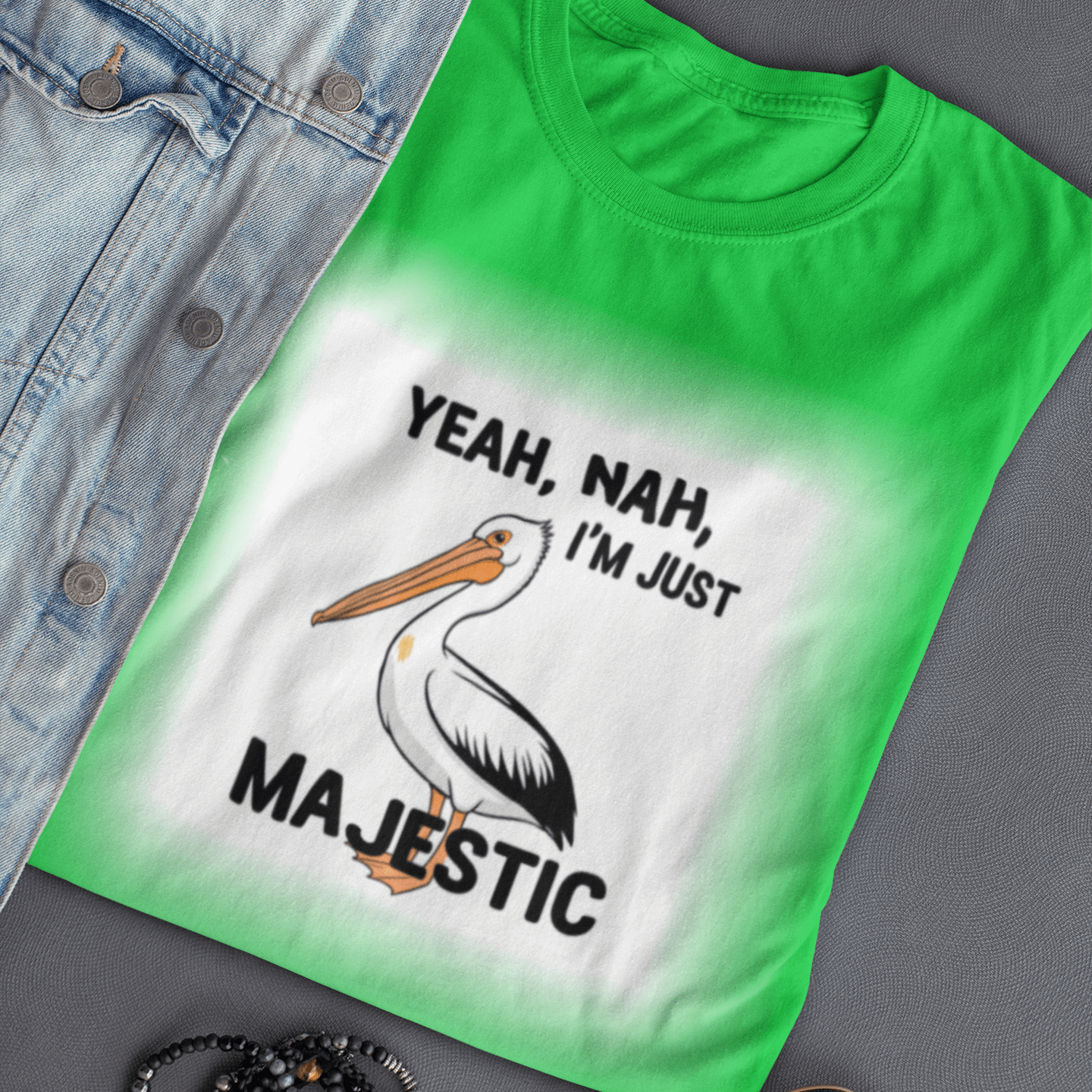 Green t-shirt with a pelican graphic and text, placed on a gray surface with sunglasses and bracelets.