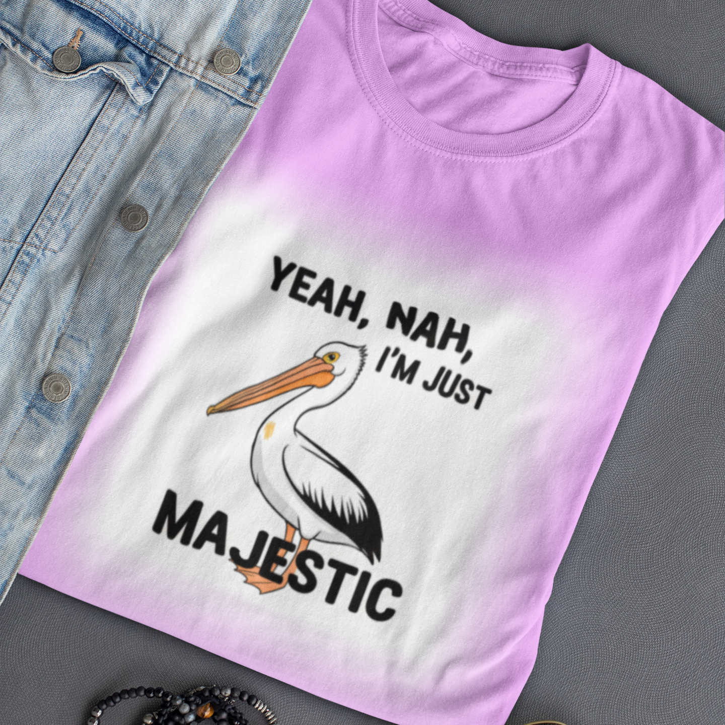 Pink t-shirt with a pelican graphic and text, placed on a gray surface with sunglasses and bracelets.