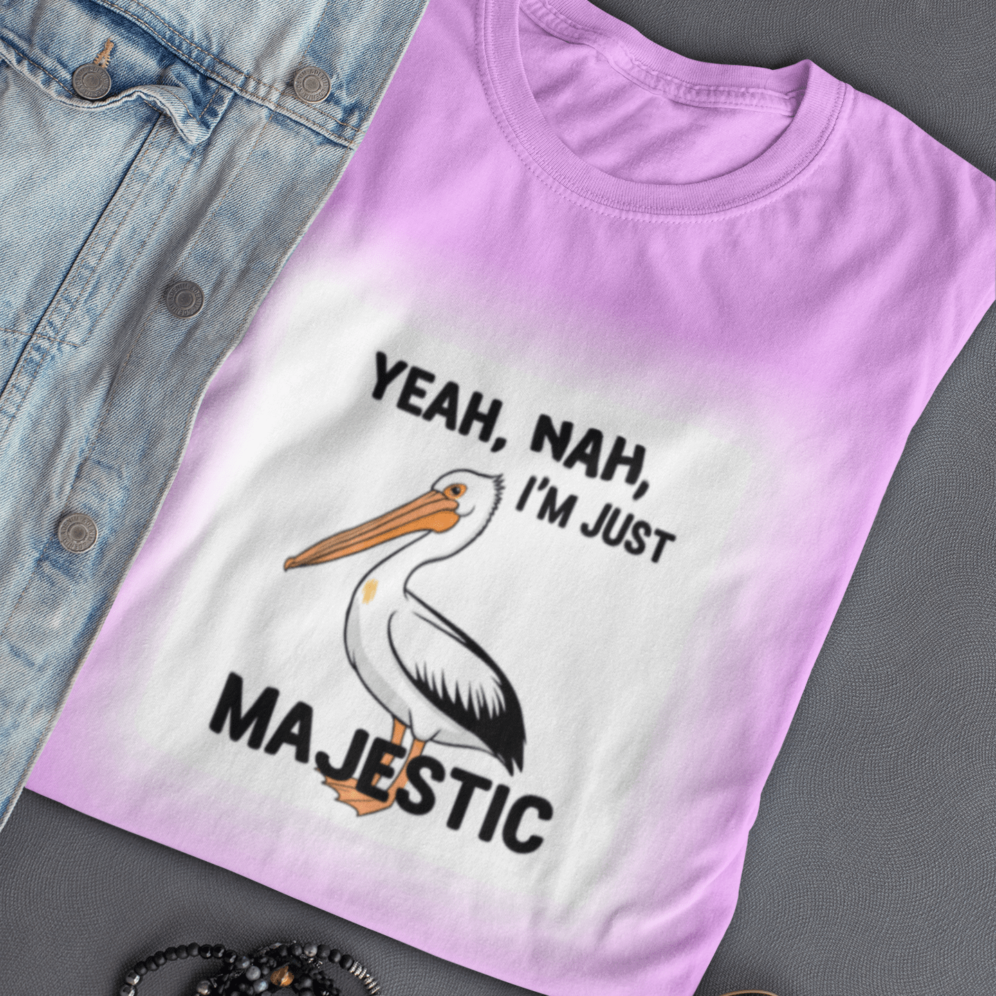 Pink t-shirt with a pelican graphic and text, placed on a gray surface with sunglasses and bracelets.