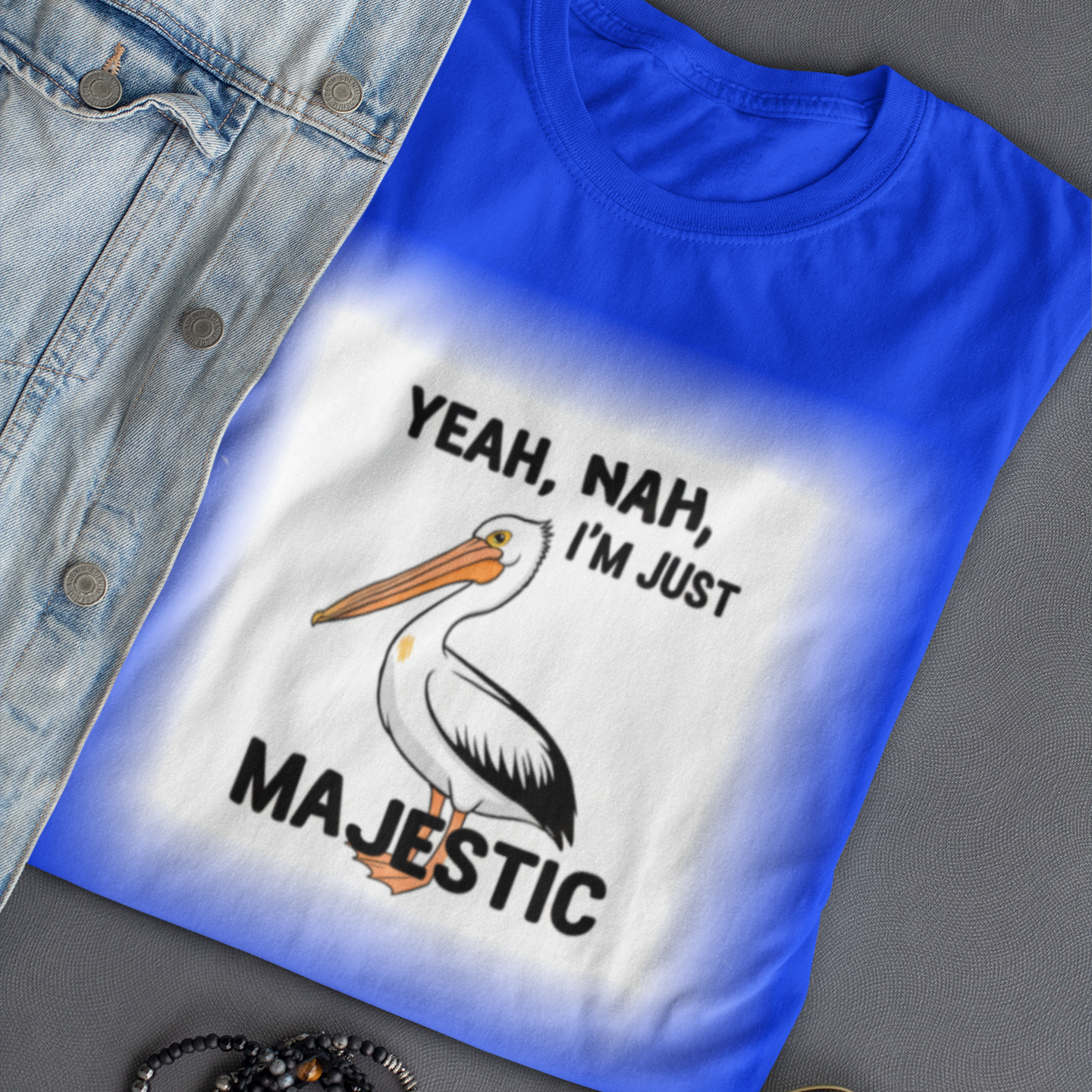 Blue t-shirt with a pelican graphic and text, placed on a gray surface with sunglasses and bracelets.