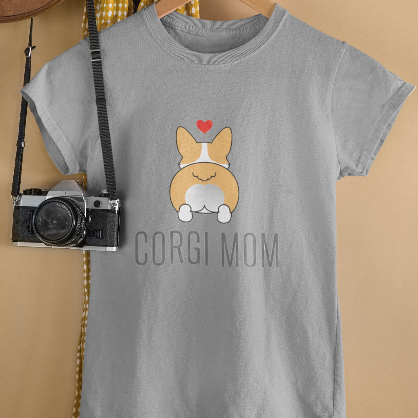 Gray t-shirt with a corgi graphic and 'Corgi Mom' text on a hanger against a beige wall.