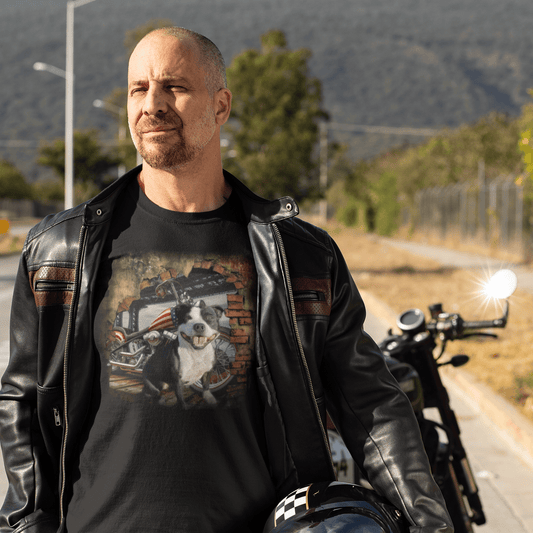 Man in a black leather jacket carrying a motorcycle helmet, wearing a black graphic t-shirt featuring a smiling Staffy dog in front of a vintage chopper motorcycle and brick wall with American flag accents.