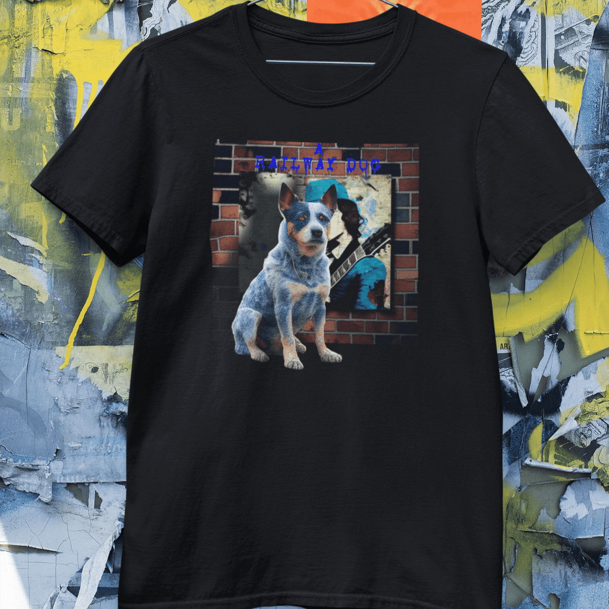 Black graphic t-shirt hanging against a distressed poster wall, featuring an Australian Cattle Dog Blue Heeler sitting in front of a brick wall mural with an electric guitar player and "A Railway Dog" text.