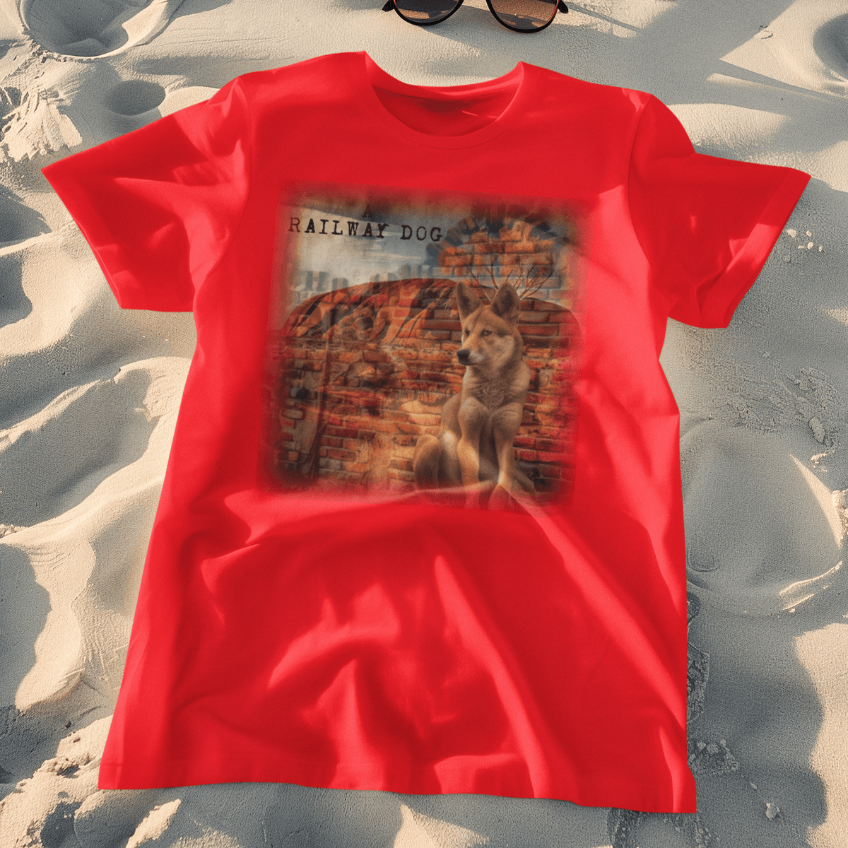 Flat-lay beach mockup of a vibrant red graphic t-shirt laid on white sand with sunglasses, featuring a wild Australian Dingo sitting in front of a brick wall mural blended with red outback rock formations and "A Railway Dog" stencil text.