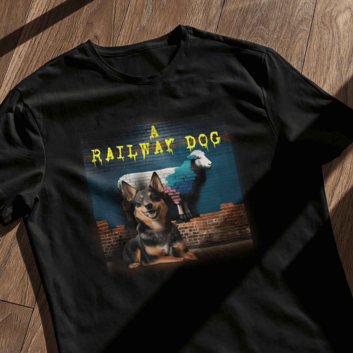 Flat-lay mockup of a black graphic t-shirt on a warm wooden floor with natural window shadows, featuring a happy Australian Kelpie dog in front of a blue brick wall with a painted Merino sheep mural and dripping yellow "A Railway Dog" text.