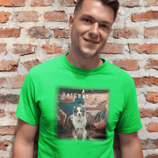 Smiling young man standing against a rustic brick wall wearing a bright green graphic t-shirt featuring an Australian Shepherd dog in front of an urban mural with a cowboy silhouette, Texas star flag design, and "A Railway Dog" text.