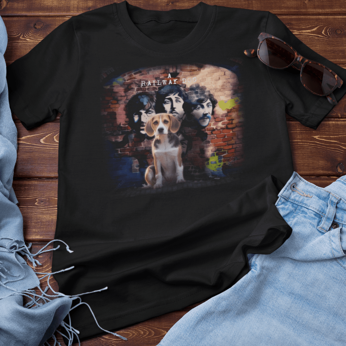 Flat-lay mockup of a black graphic t-shirt arranged on rustic wood planks next to denim jeans and sunglasses, featuring a Beagle puppy sitting in front of a brick wall mural depicting vintage 1960s British rock band portraits with "A Railway Dog" text.