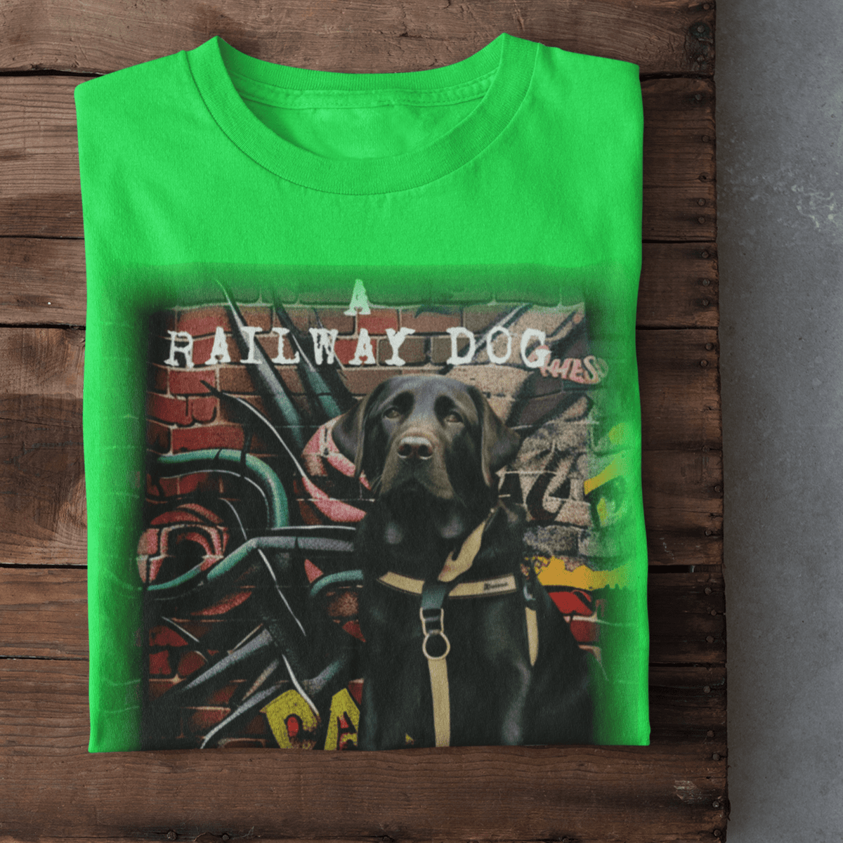 Folded bright neon green graphic t-shirt laid on rustic dark wood planks, featuring a Black Labrador Retriever wearing a harness in front of a colorful graffiti brick wall with "A Railway Dog" stencil text. T-shirt featuring a Labrador Retriever working as a guide dog.