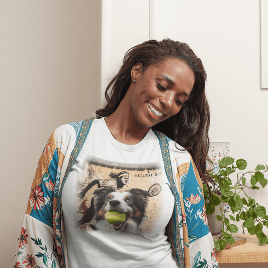 Smiling woman at home wearing a white graphic t-shirt featuring an Border Collie catching a tennis ball in front of street art of a retro tennis player and "A Railway Dog" text.