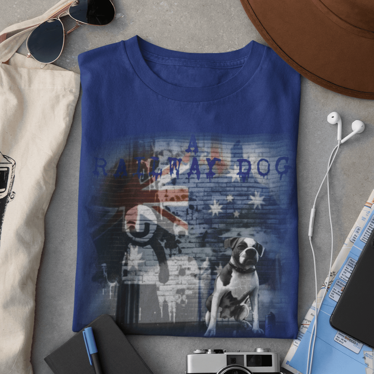 Flat-lay travel accessories mockup featuring a royal blue graphic t-shirt with a Boxer dog seated before an urban brick wall mural of the Australian flag with blue dripping "A Railway Dog" text.