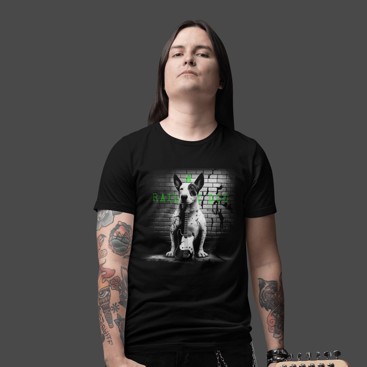 Long-haired man with arm tattoos wearing a black graphic t-shirt featuring a black and white Bull Terrier sitting with a bass guitar in front of a brick wall with green "A Railway Dog" text.