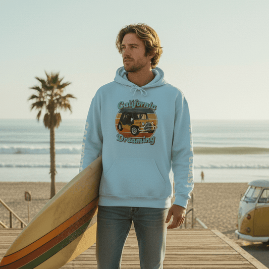 Man wearing a California Dreaming West Coast Hoodie holding a surfboard on a wooden boardwalk with palm trees and a van in the background.