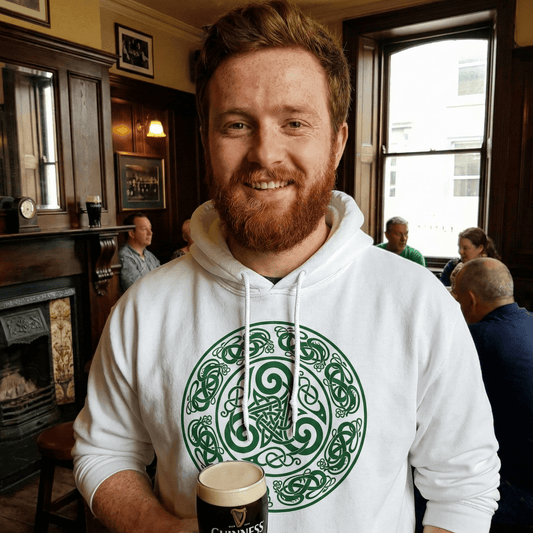 Man in a white hoodie with a green Celtic design holding a Guinness beer mug in a pub setting.