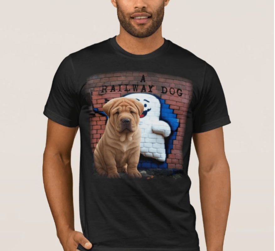 Man modeling a black graphic t-shirt featuring a wrinkled Shar-Pei puppy sitting in front of a brick wall cutout revealing a retro cartoon ghost mascot mural with "A Railway Dog" stencil text.