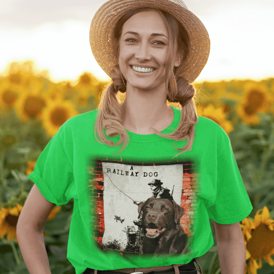 Smiling woman in a straw hat standing in a sunflower field wearing a bright green t-shirt featuring a Chocolate Labrador retriever in front of street art depicting a vintage fisherman and "A Railway Dog" poster on a brick wall.