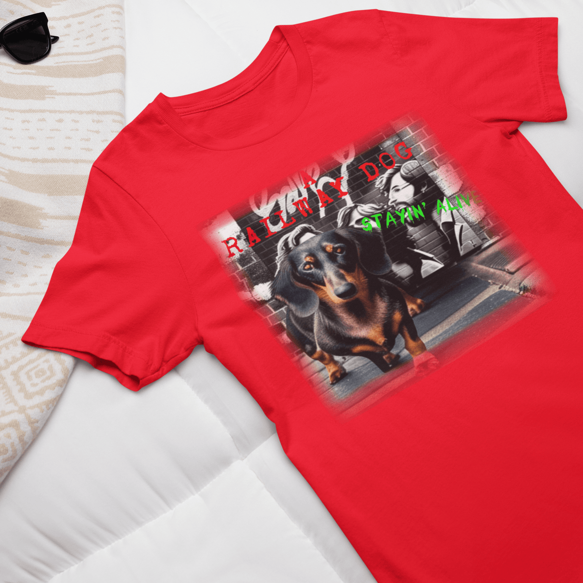 Flat-lay travel outfit mockup of a bright red graphic t-shirt laid on a white duvet next to sunglasses, featuring a black and tan Dachshund dog in front of a brick wall mural with disco street art and "A Railway Dog Stayin' Alive" text.
