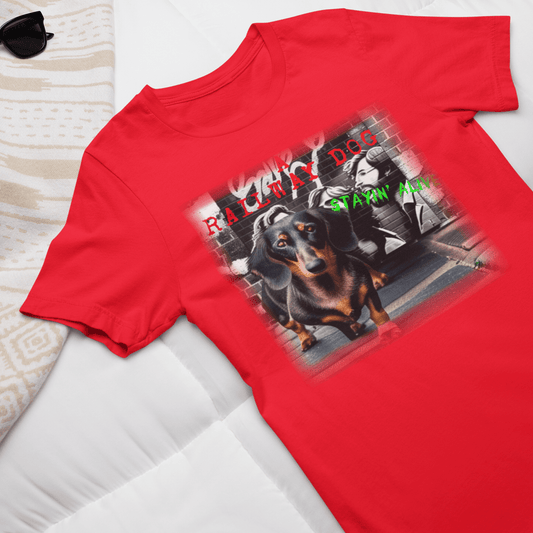 Flat-lay travel outfit mockup of a bright red graphic t-shirt laid on a white duvet next to sunglasses, featuring a black and tan Dachshund dog in front of a brick wall mural with disco street art and "A Railway Dog Stayin' Alive" text.