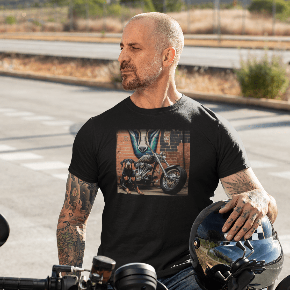 Man with arm tattoos sitting on a motorcycle holding a helmet, wearing a black graphic t-shirt featuring a Doberman Pinscher sitting beside a custom chopper motorcycle in front of an urban brick wall mural.
