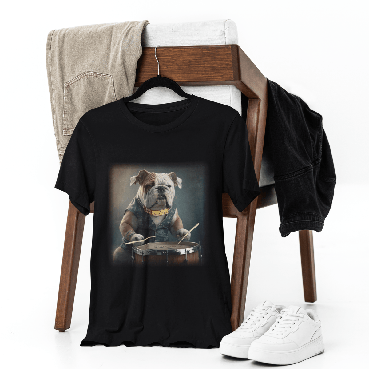 Black graphic t-shirt displayed on a hanger on a wooden chair with white sneakers and jeans, featuring an English Bulldog wearing a denim vest playing a snare drum with drumsticks.