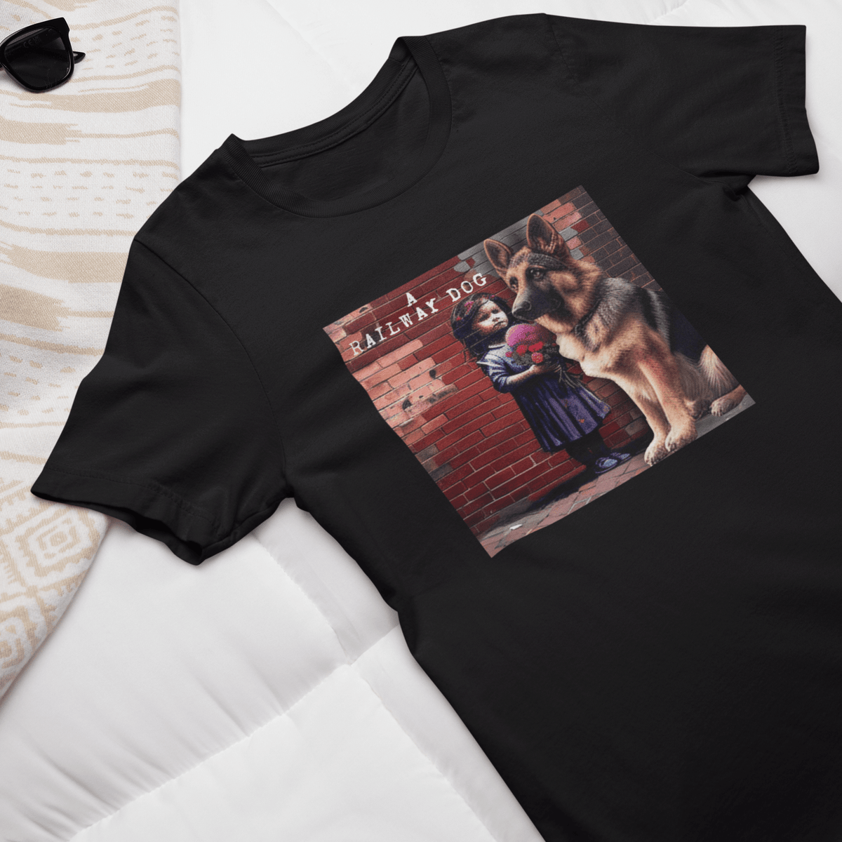 Flat-lay display of a black graphic t-shirt laid on a white bed with sunglasses, featuring a German Shepherd sitting beside a street art stencil of a young girl holding flowers against a brick wall with "A Railway Dog" text.