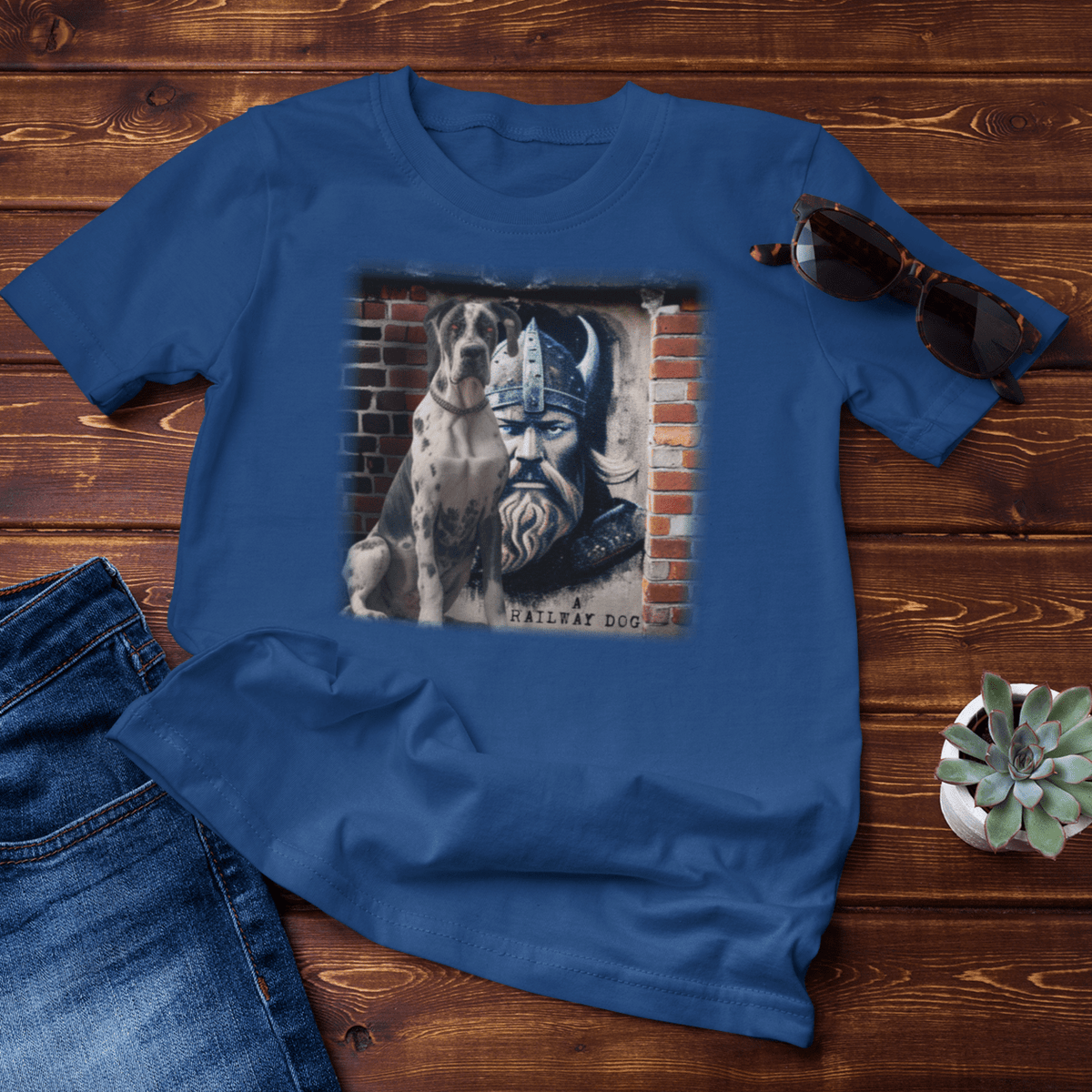 Flat-lay mockup on dark wood boards featuring a royal blue graphic t-shirt styled with blue jeans, sunglasses, and a potted succulent, printed with a Harlequin Great Dane sitting beside an urban brick mural of a horned Viking warrior with "A Railway Dog" text.