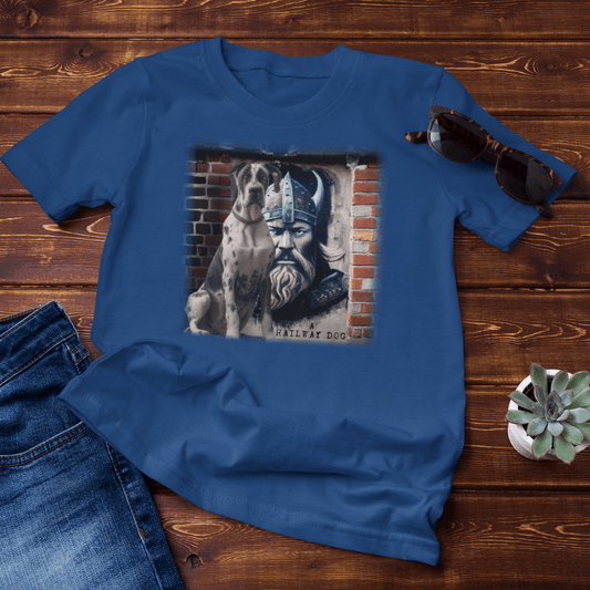 Flat-lay mockup on dark wood boards featuring a royal blue graphic t-shirt styled with blue jeans, sunglasses, and a potted succulent, printed with a Harlequin Great Dane sitting beside an urban brick mural of a horned Viking warrior with "A Railway Dog" text.