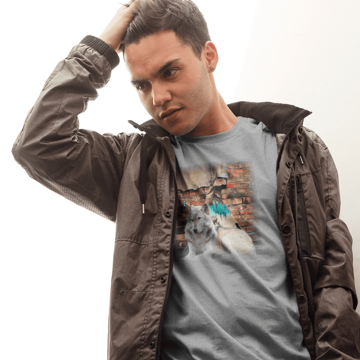 Young man in an open jacket wearing a heather grey graphic t-shirt featuring a grey wolf portrait against a distressed brick wall with teal street art accents.