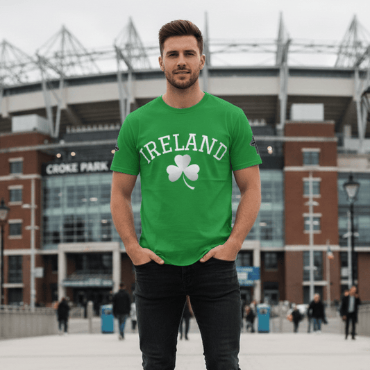 man stands in the foreground of this full-length shot, wearing a bright green T-shirt with the word "IRELAND" and a white shamrock printed on the front. He is also wearing dark blue jeans and brown leather boots. Behind him is the Croke Park stadium in Dublin, Ireland. The man is smiling and has his hands in his pockets. The sky is overcast and the ground is paved with light-colored stones. There are a few other people walking in the background.