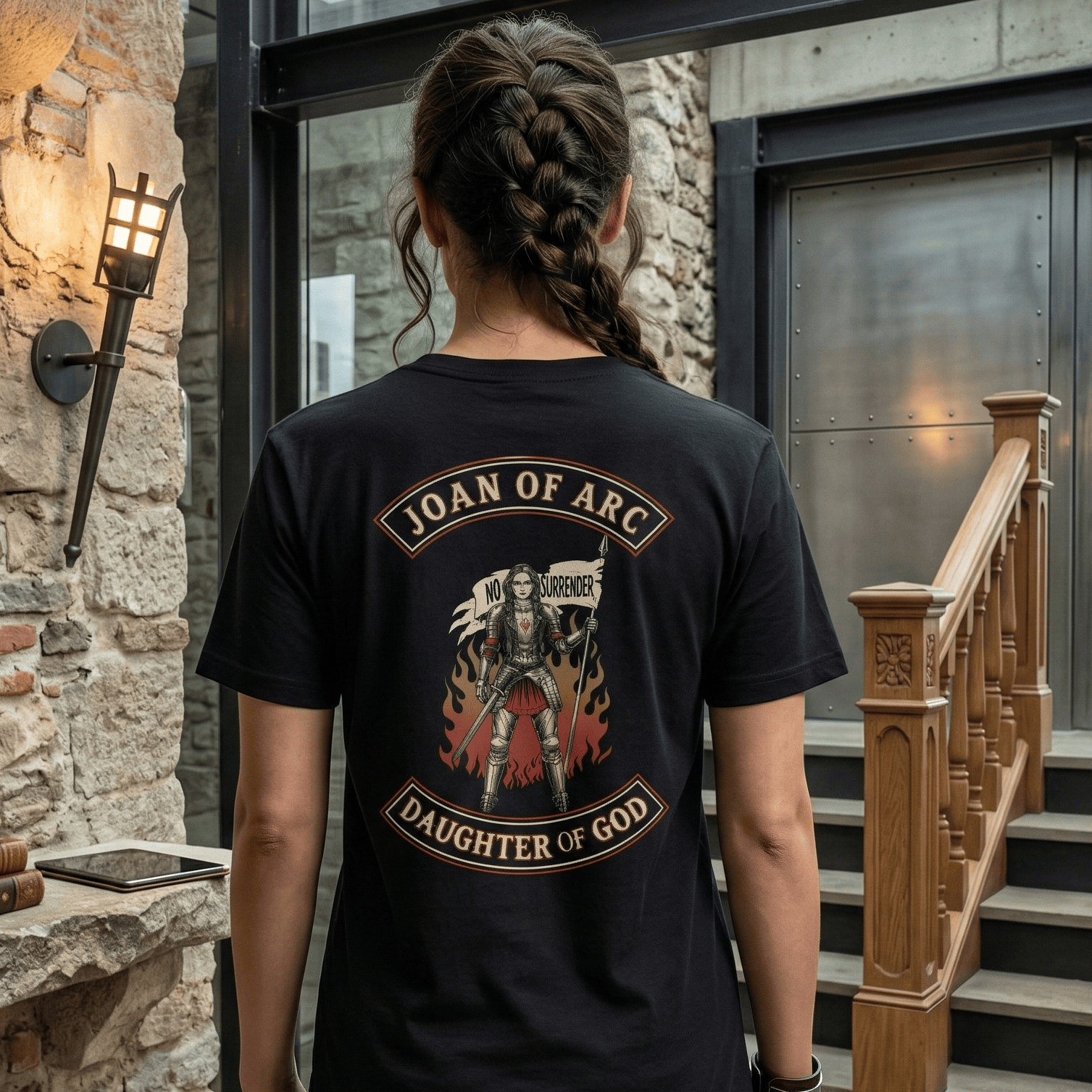 An image of the back of a woman model wearing a black t-shirt. On the back of the t-shirt is a graphic featuring Joan of Arc, depicted as a medieval knight in armor, holding a sword and a banner that reads "NO SURRENDER." The graphic is framed by two arched banners: the top one says "JOAN OF ARC" and the bottom one says "DAUGHTER OF GOD." The woman's hair is in a long braid. She is standing in a modern building with medieval elements, such as a stone wall, a metal helmet on a shelf, and a modern staircase.