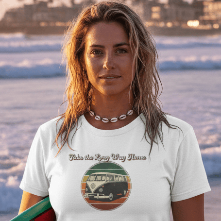 Woman wearing a white VW Kombi tee holding a colorful surfboard on a beach with waves and palm trees in the background