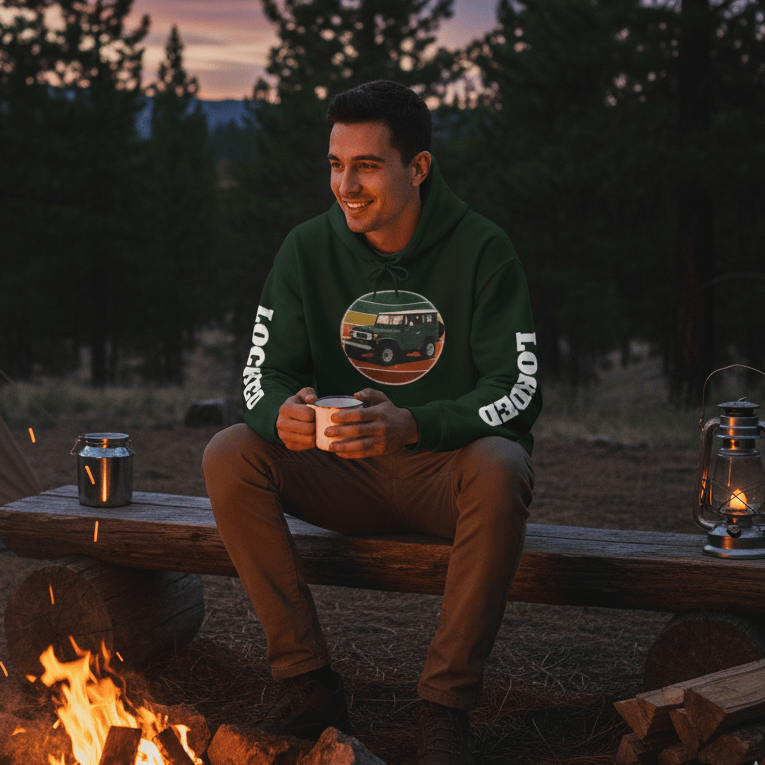 Man sitting by a campfire in a forest at dusk, wearing a green sweatshirt with a graphic design.