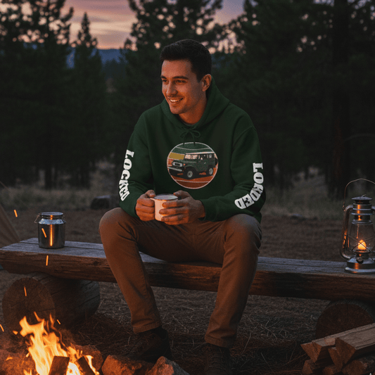 Man sitting by a campfire in a forest at dusk, wearing a green sweatshirt with a graphic design.