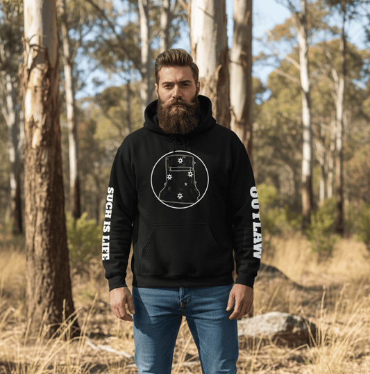 A full-length shot of a bearded man standing in a sun-drenched Australian bushland setting. He is wearing a black hoodie featuring a white circular graphic of Ned Kelly’s iconic armor helmet, which is accented with stars from the Southern Cross. The sleeves of the hoodie are printed with vertical white text: "SUCH IS LIFE" on the right arm and "OUTLAW" on the left. He is also wearing blue jeans and brown leather work boots. The background consists of tall eucalyptus trees and dry, grassy ground.