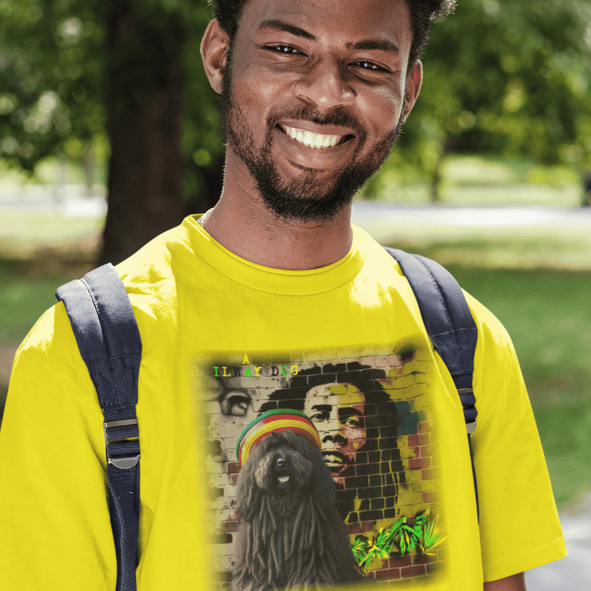 Smiling young man outdoors with a backpack wearing a bright yellow t-shirt featuring a dreadlocked Puli dog wearing a Rasta tam hat in front of a Bob Marley brick wall mural with "A Railway Dog" text.