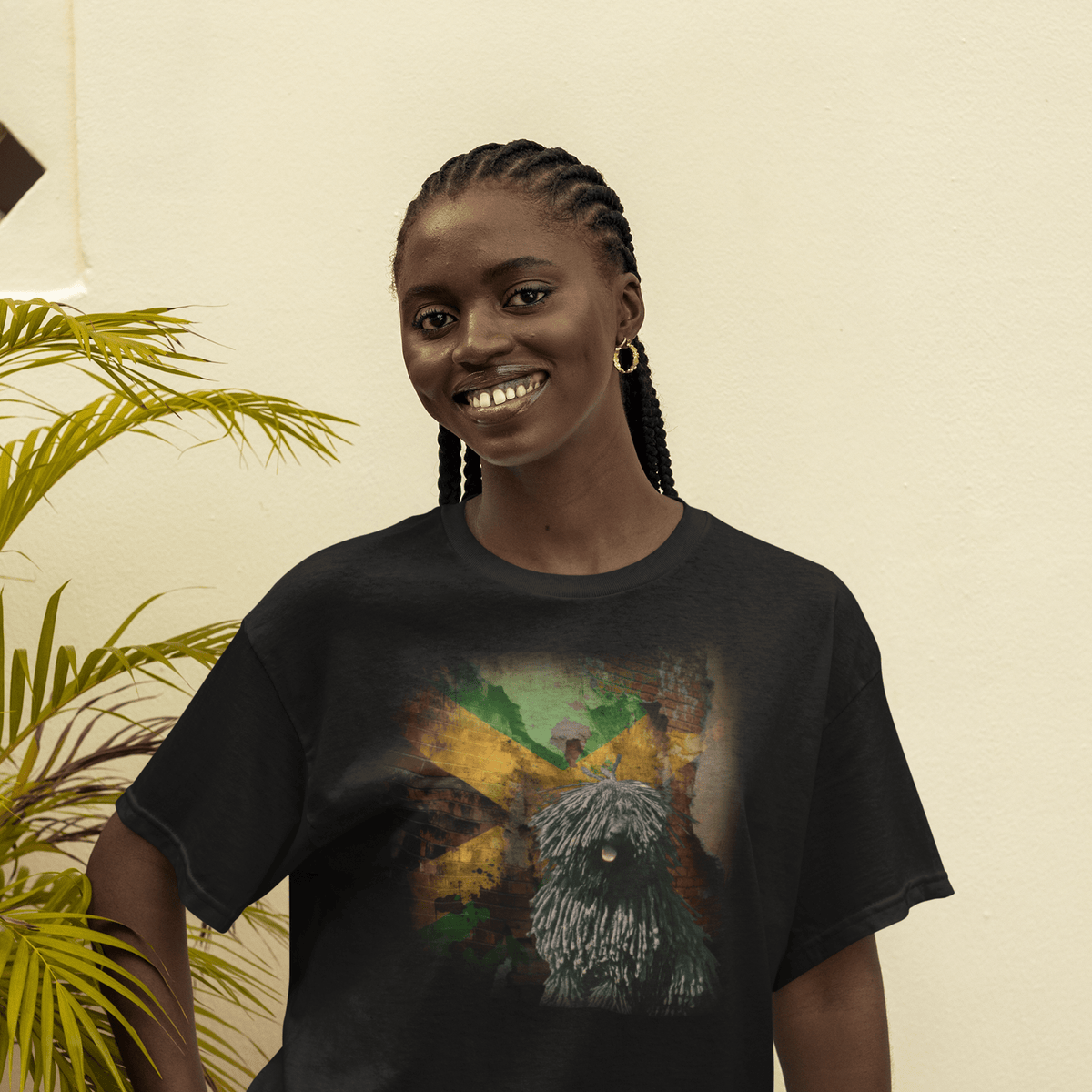 Smiling woman posing outdoors in a black graphic t-shirt featuring a corded black Puli dog in front of a distressed brick wall painted with the Jamaican flag.