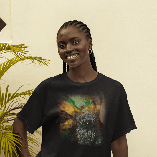 Smiling woman posing outdoors in a black graphic t-shirt featuring a corded black Puli dog in front of a distressed brick wall painted with the Jamaican flag.