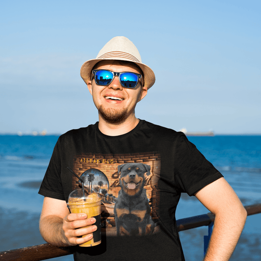 Smiling man in sunglasses and a straw hat holding an iced drink while wearing a black graphic t-shirt featuring a happy Rottweiler dog in front of a brick wall with palm trees and "A Railway Dog" text.