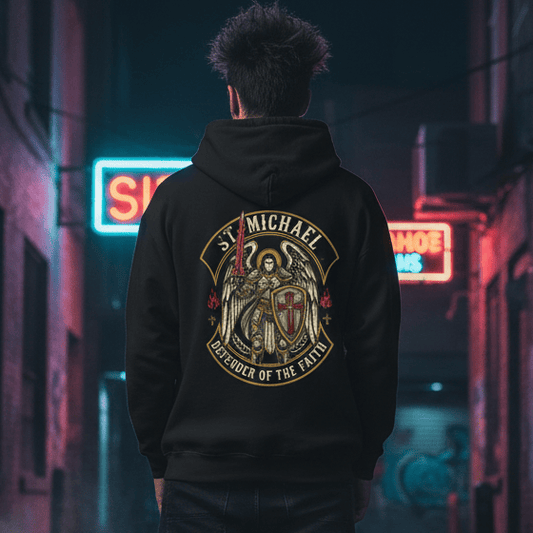 The image captures a model from the back, standing in a dimly lit, narrow urban alleyway at night. The model wears a black hoodie featuring a large, detailed graphic on the back of St. Michael in armor with wings, holding a flaming sword and a shield, surrounded by the text "ST. MICHAEL DEFENDER OF THE FAITH."

The setting is gritty and atmospheric, with damp pavement reflecting the vibrant blue and pink glow of neon signs in the background. Wisps of fog or steam rise from the ground, adding to