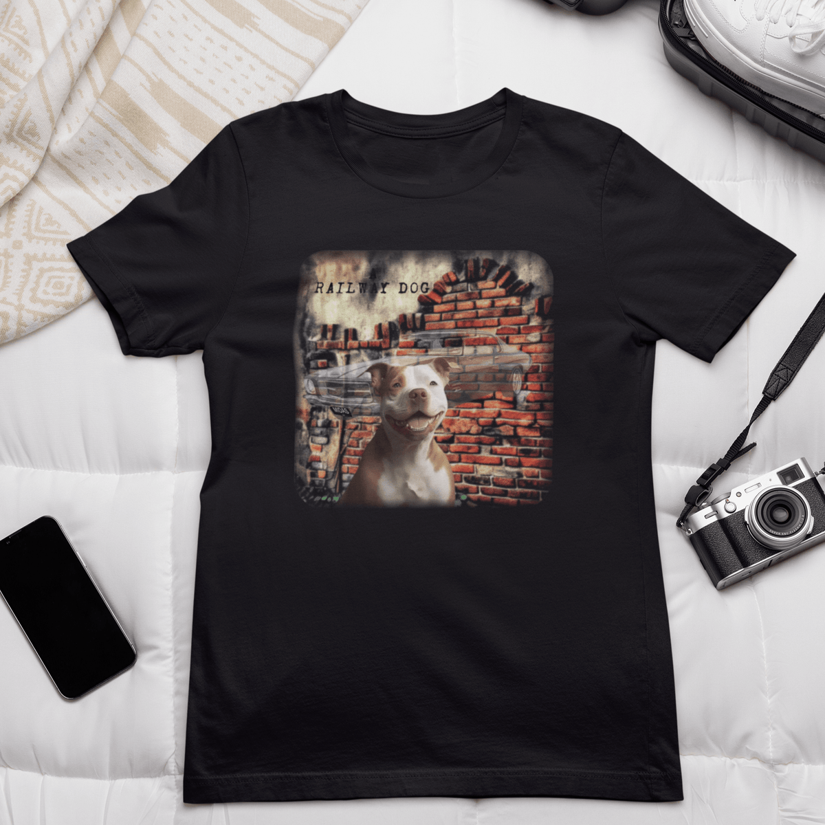 Flat-lay bed mockup featuring a black graphic t-shirt styled next to a vintage camera, smartphone, and white sneakers, printed with a smiling brown and white Staffy in front of a classic Holden Monaro  mural and exposed brick wall with "A Railway Dog" text. Staffy HQ T-Shirt | Staffordshire Bull Terrier HQ Tee | That Should Be on a T-Shirt