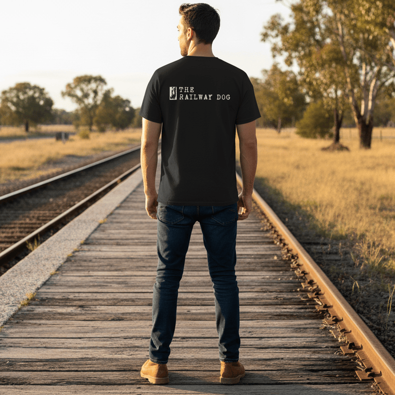 Man standing on a wooden platform by train tracks in a rural setting with 'The Railway Dog' t-shirt.