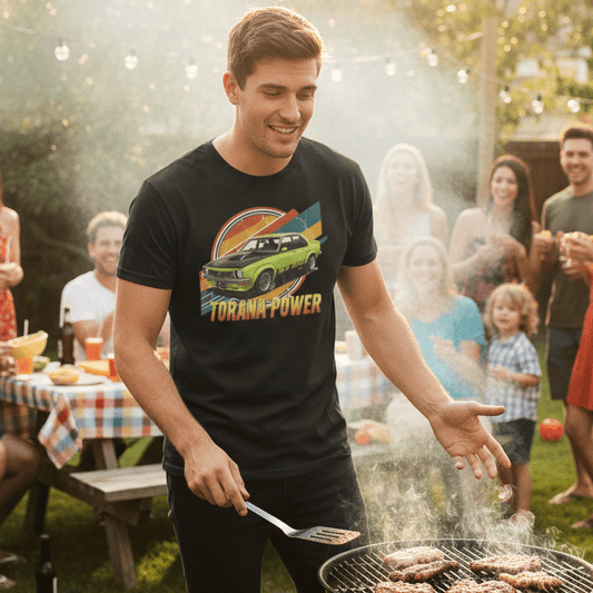 Man wearing a Holden Torana Tshirt grilling food at a backyard barbecue with friends and family in the background